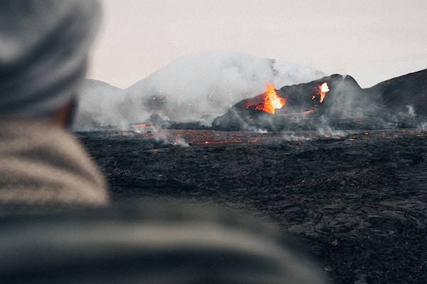 Un homme de dos devant un volcan en éruption, avec de la lave qui jailli et des fumées.