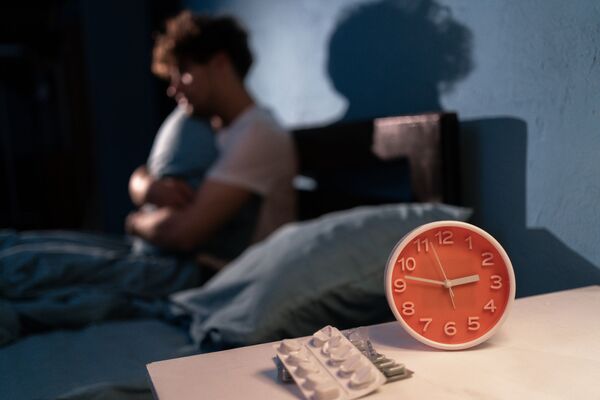 un jeune homme dans son lit incapable de dormir. devant lui un réveil et des médicaments posés sur une table.